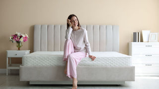 Woman in pajamas sitting on modern upholstered bed with white mattress in bright bedroom