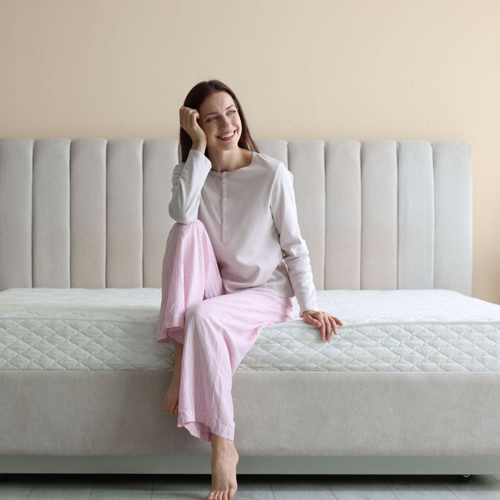 Woman in pajamas sitting on modern upholstered bed with white mattress in bright bedroom