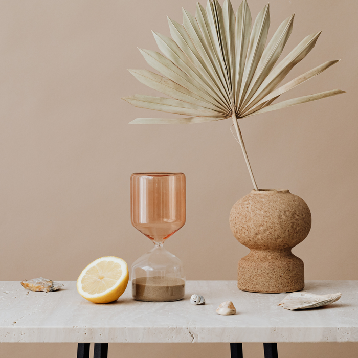 Modern hourglass sand timer with seashells, lemon slice, ceramic vase, and palm leaf on marble table