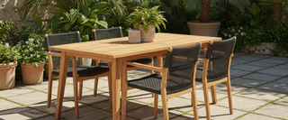 Outdoor wooden dining table with four black woven chairs on a patio, surrounded by potted plants.