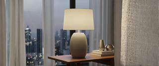 Modern table lamp with beige shade on wooden nightstand by window, city view at dusk