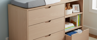 Light wood nursery dresser with changing pad, storage shelves, baby clothes, and books