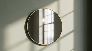 Modern round wall mirror with gold frame reflecting window light on pale wall
