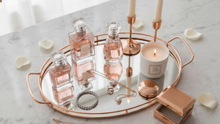 Rose gold vanity tray with perfume bottles, candle, jewelry, and petals on marble table