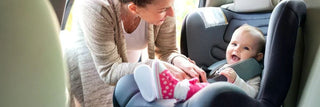 Smiling baby secured in a car seat with a woman adjusting the harness inside a vehicle.