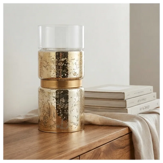 A two-tiered gold-toned candle holder with a clear glass top, styled on a wooden table next to a stack of books and a draped beige cloth.