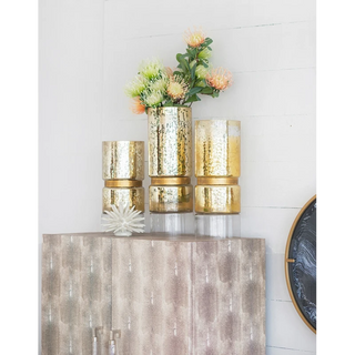 Three metallic gold  textured cylindrical vases - candle holders arranged on a mirrored cabinet, with artificial flowers in the tallest vase, against a white paneled wall.
