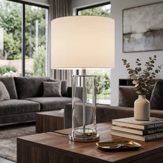 A modern table lamp with a large white drum shade and a clear cylindrical glass base with a polished silver metal trim, shown lit on a wooden coffee table in a contemporary living room setting.