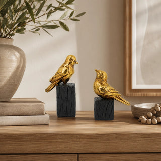 Two decorative gold bird figurines perched on dark textured pedestals, displayed on a wooden console table in a styled interior setting with a vase, books, and a bowl.