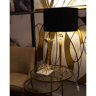 Tall gold table lamp with black shade styled beside armchair on glass side table – modern living room lighting, Shopica.
