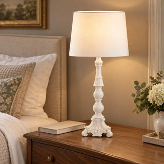 Ornate candlestick lamp glowing on wooden bedside table beside linen headboard